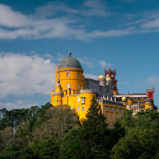The vibrant Pena Palace in Sintra Portugal with its yellow facade and red clock tower, surrounded by green trees O vibrante Palácio da Pena em Sintra Portugal com as suas fachadas amarelas e telhado vermelho, rodeado por árvores verdes