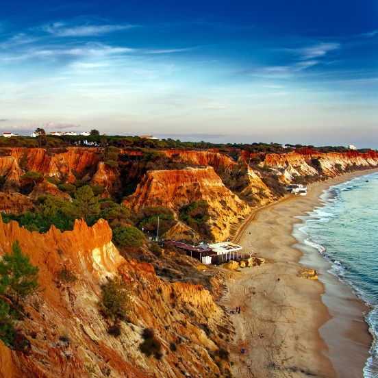 The iconic red cliffs of the Algarve coastline overlooking a golden sandy beach and the Atlantic Ocean. As icónicas falésias vermelhas da costa do Algarve com vista para uma praia de areia dourada e o Oceano Atlântico.