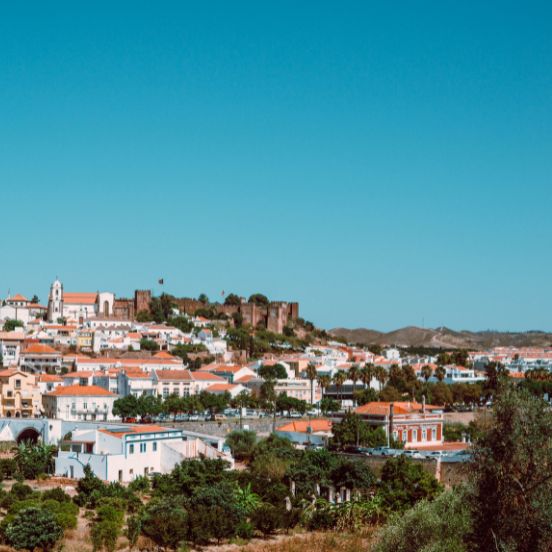 A panoramic view of the historic town of Silves with its red-brick castle on the hilltop and white buildings. Uma vista panorâmica da cidade histórica de Silves com o seu castelo de tijolos vermelhos no topo da colina.