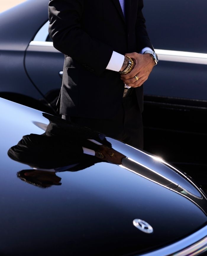 English: A professional chauffeur in a suit standing next to a black Mercedes-Benz luxury car Português: Um motorista profissional de fato e gravata em pé junto a um carro de luxo preto da Mercedes-Benz