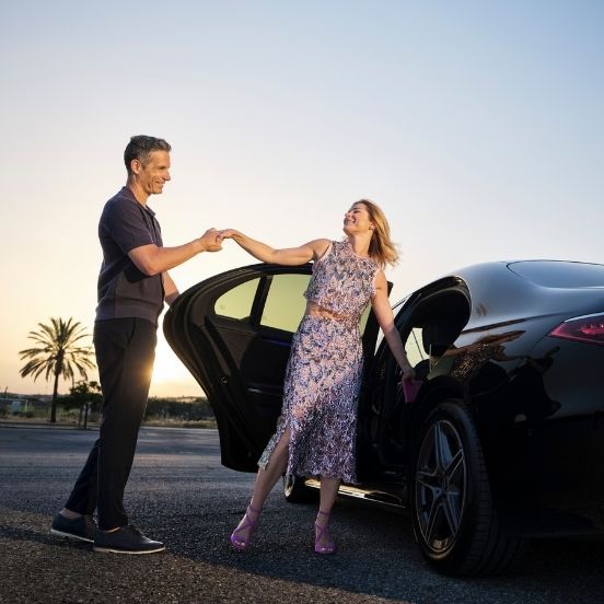 English: A man helping a woman out of a black luxury car at sunset, representing a chauffeur service for a special event or a private happy airport transfer Português (Europeu): Um homem a ajudar uma mulher a sair de um carro de luxo preto ao pôr do sol, representando o serviço de transporte para um evento ou um transfer de aeroporto alegre