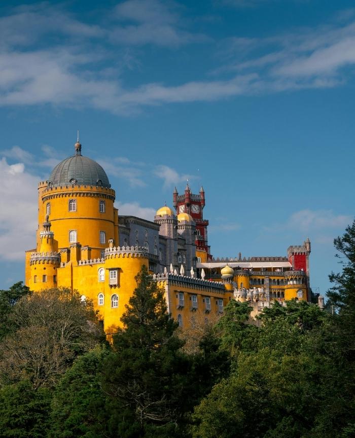 The vibrant Pena Palace in Sintra Portugal with its yellow facade and red clock tower, surrounded by green trees O vibrante Palácio da Pena em Sintra Portugal com as suas fachadas amarelas e telhado vermelho, rodeado por árvores verdes