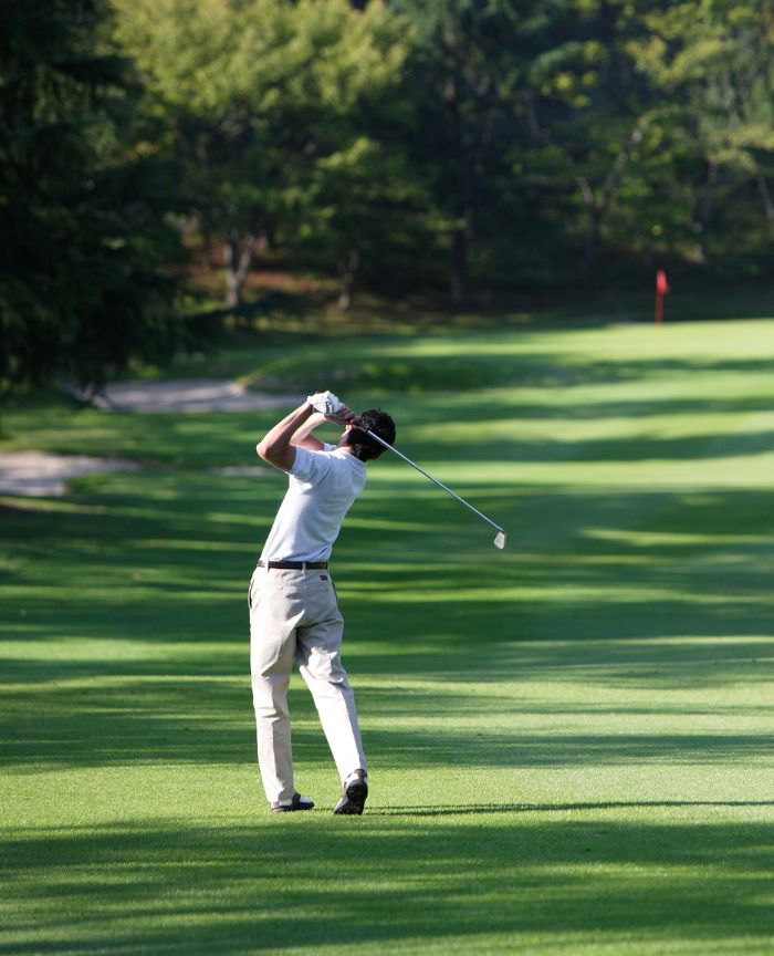 English: A golfer in a white shirt swinging a golf club on a lush green fairway with a red flag in the distance Português: Um golfista de t-shirt branca a fazer um swing num green relvado com uma bandeira vermelha ao fundo
